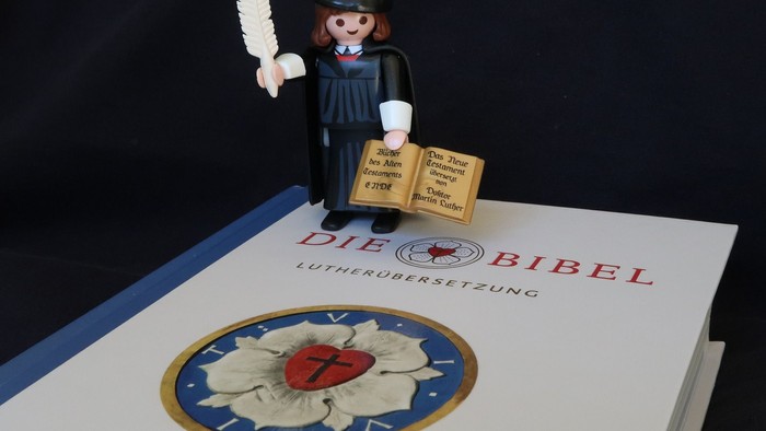 Eine Playmobil-Figur hält ein Buch und steht auf einem Buch mit einem blauen Einband und einem roten Logo.
