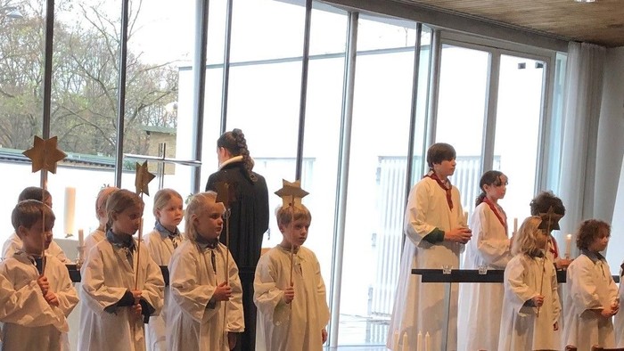 En gruppe børn i hvide kapper og sort stander i en rum med store vinduer og læreren står foran dem