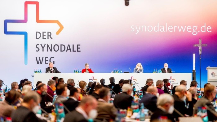 Eine große Versammlung mit Menschen, die an Tischen sitzen, vor einem Podium mit Rednern und einem großen Kreuz-Symbol im Hintergrund.