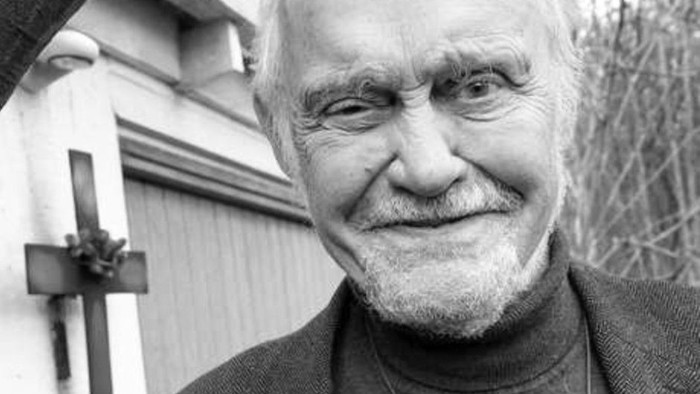 En eldre mand med kvæde skæg står udenfor et hus med træmur.