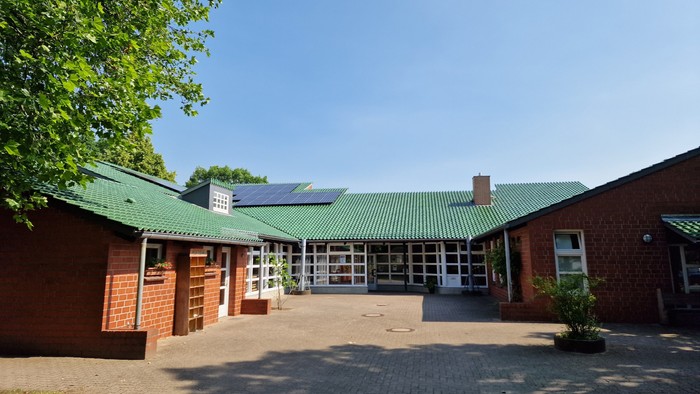 Das Bild zeigt ein rotes Backsteingebäude mit grünem Dach und Solarpanels, umgeben von Bäumen und einem gepflasterten Hof.