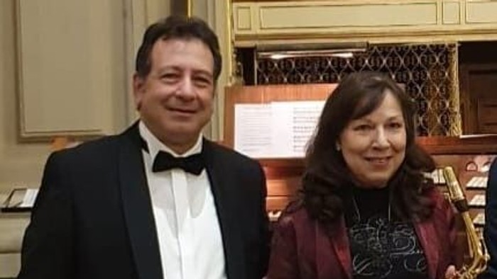 Zwei Personen posieren mit einem Mann, der ein Saxhorn hält, neben einer verzierten Pfeifenorgel in einem formellen Rahmen.