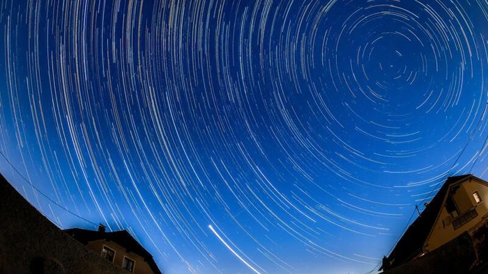 Atemberaubender Nachthimmel mit faszinierenden Sternenspuren und einer schwachen Meteorspur über einem Dorf.