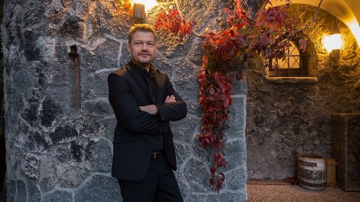 Mann in dunklem Anzug steht vor steinerner Wand mit Weinreben.