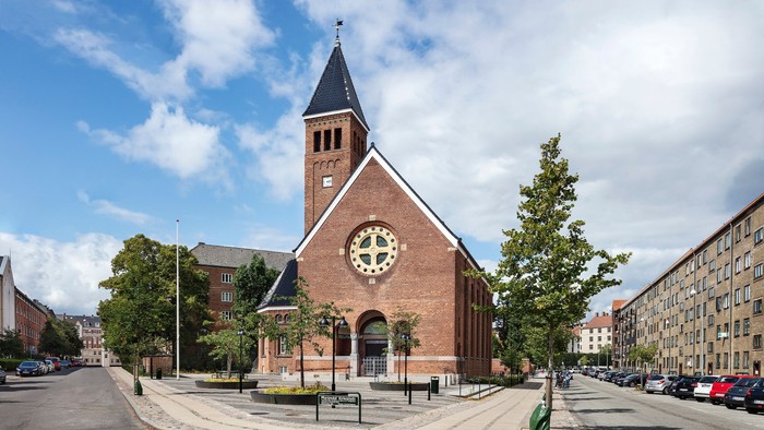Murstenskirke med højt spir og ur, omgivet af træer og bygninger.