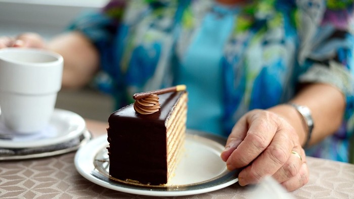 Eine Person hält einen Teller mit einem Stück Schokoladenkuchen und einer Tasse Kaffee steht daneben