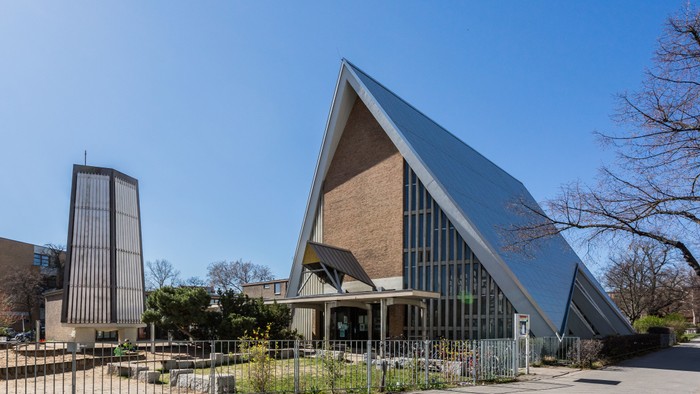 Modernes Gebäude mit markanter dreieckiger Architektur und Glasfassade, umgeben von Bäumen und einem Zaun.