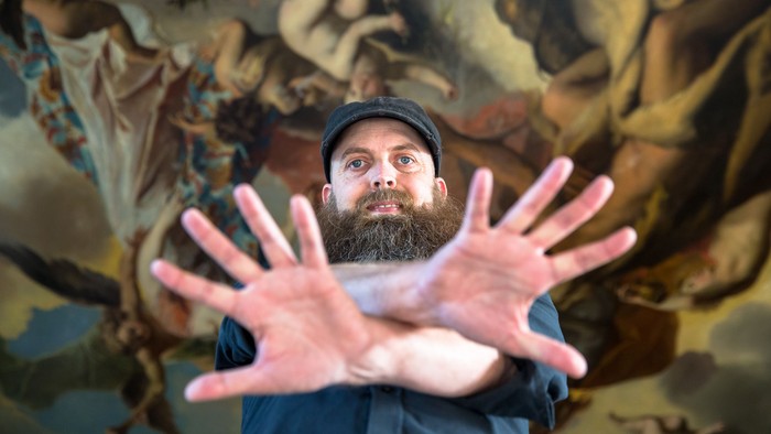 Mand med skæg og mørk behøjning har hænderne op foran maleri