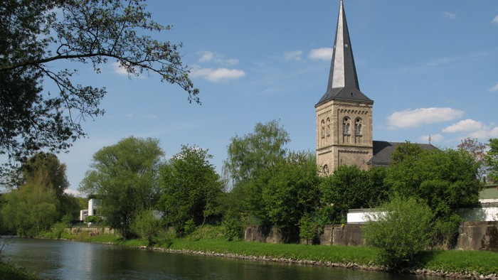 Ein hoher Kirchturm mit spitzer Spitze steht neben einem Fluss, umgeben von üppigen grünen Bäumen unter einem klaren blauen Himmel