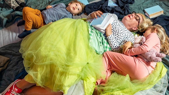 To børn ligger på sengen med klær og en gul pukkelpude.