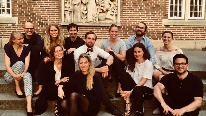 En gruppe af unge mænd og kvinder sidder på trin i forgrunden af et murstenshus med en relief.