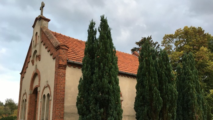 Kleine Kirche mit rotem Dach und drei hohen grünen Bäumen davor.