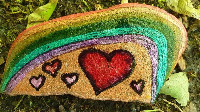 Bemalter Stein mit Regenbogenstreifen, Herz und kleineren Herzen auf einer natürlichen Oberfläche.