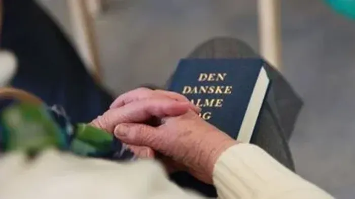 En person holder en blå bog med tittelen "Den Danske Alme Bog".