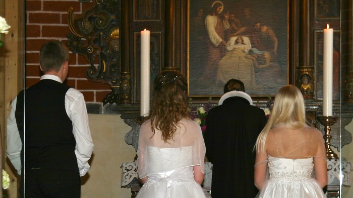 En familie i kirke, to piger i hvide spidsdresser og en dreng i blå, foran alteret med lysstake.