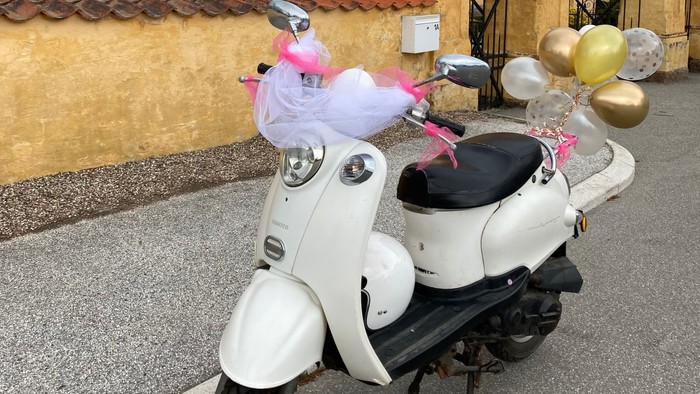 En hvid scooter med balloner og tøj på bagageboksen, parkeret foran et slotslignende hus