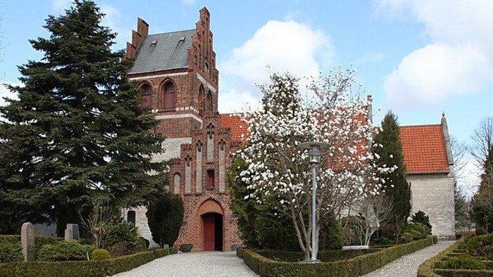 Et gammelt kirkebygning med røde murværker og spisse tårn, omgivet af træer og buske.