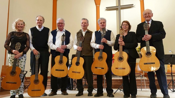 Eine Gruppe von sieben Personen steht auf einer Bühne und hält Gitarren.