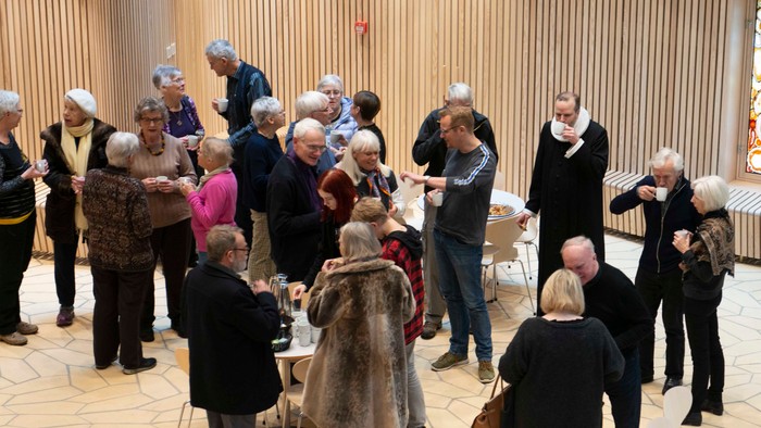En gruppe ældre mennesker samles i et rum med høje kolonner og drikker kaffe.