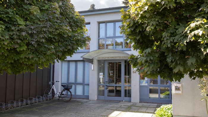 Das Bild zeigt ein weißes Gebäude mit Säulen und einem Vordach, umgeben von Bäumen und einem Zaun.