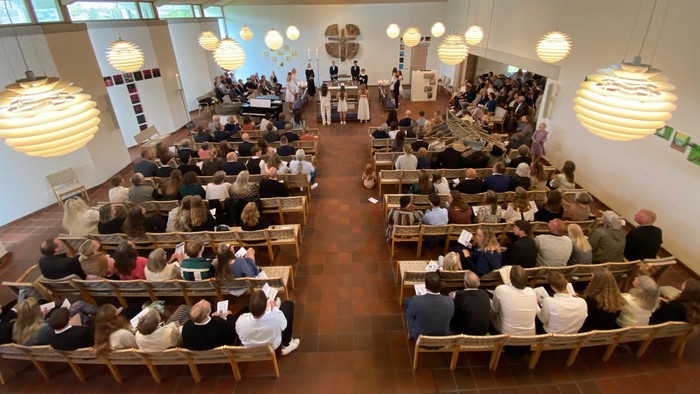 En stor hall med mange folk, der sidder på træbænke og ser mod en scene.