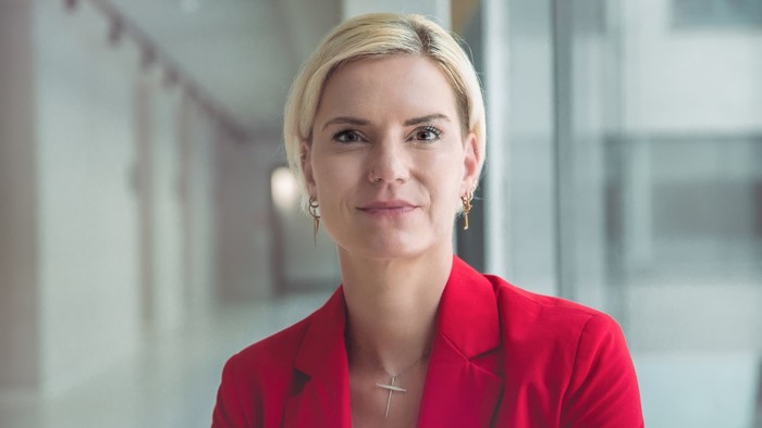 Eine Frau mit blondem Haar in einem roten Blazer posiert selbstbewusst.