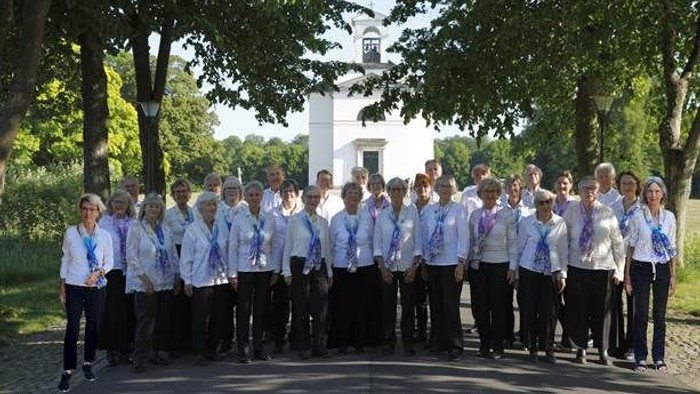 En gruppe korister poser i forgrunden af et hvidt tårn med klokke.