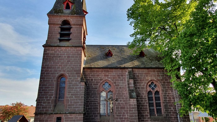 Kirchengebäude mit hohem Turm und spitzem Dach, umgeben von einem Holzzaun und Bäumen