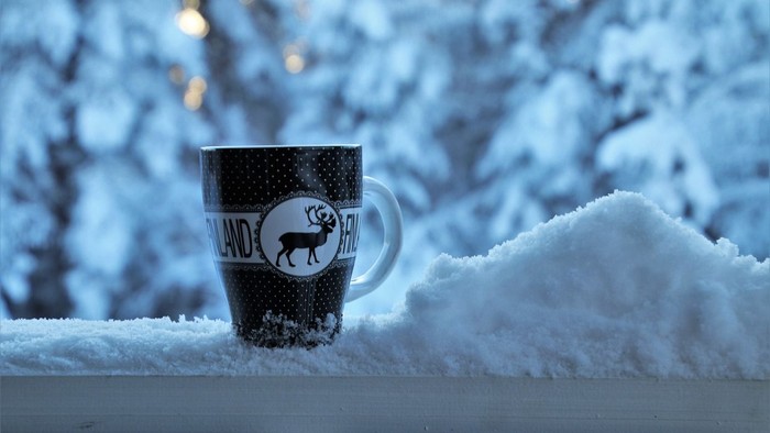 Eine Tasse mit Rentier-Dekor steht im Schnee auf einer weißen Oberfläche vor einem schneebedeckten Baum.