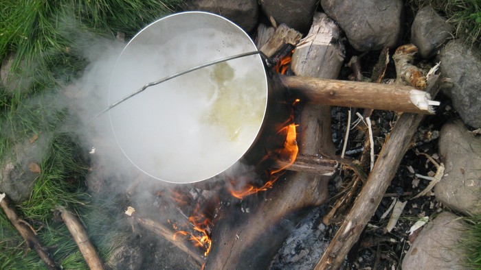 Ein Topf mit dampfendem Inhalt steht über einem Lagerfeuer im Freien.