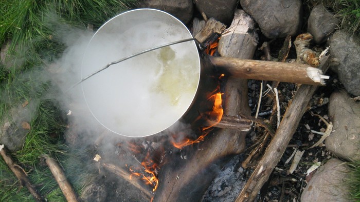 Ein Topf mit dampfendem Inhalt steht über einem Lagerfeuer im Freien.