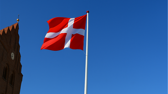 Et dansk flag med et hvidt kors på rød baggrund vajer tydeligt mod en klar blå himmel.
