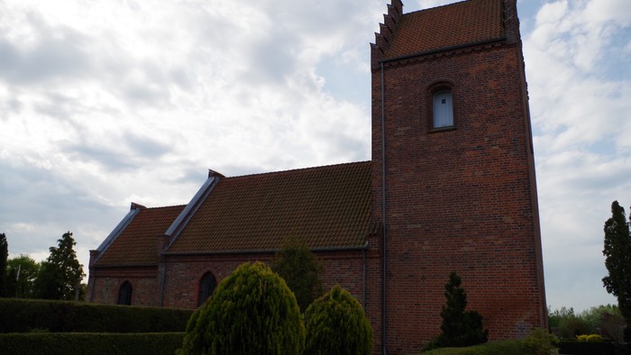 En rød murstenskirke med tårn og smukke træer foran