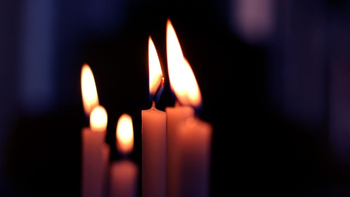 Tre tændte stearinlys på rad, med flammerne flakkende, placeret mod en mørk baggrund.