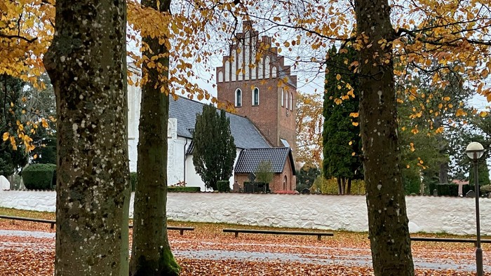 En kirke med tårn og spiret, omgivet af faldne løv og træer i efterårsfarver