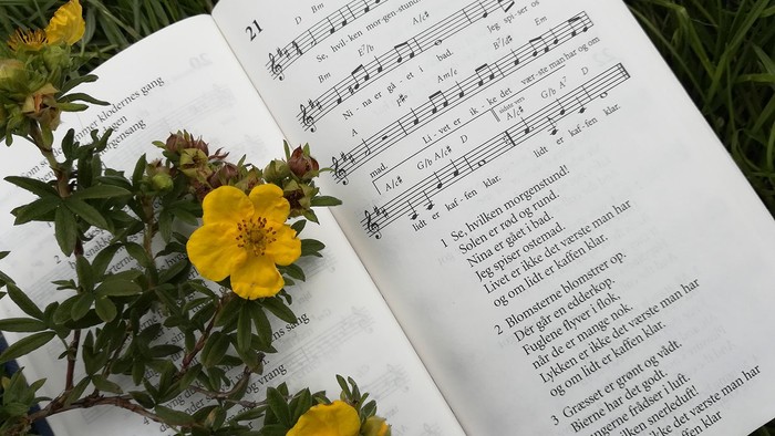 En bog med noter og tekster ligger på græs med gule blomster der er lagt på den.