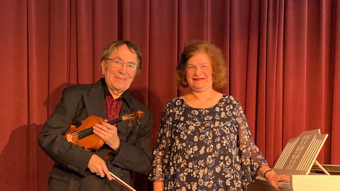 Ein Mann und eine Frau posieren vor einem roten Vorhang, der Mann hält eine Geige.