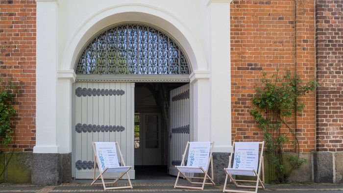 Vor einem weißen Tor stehen drei Stühle mit Schildern.