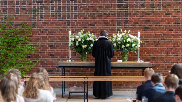 En præst står ved alteret foran murstenge væg med blomsterarrangementer.