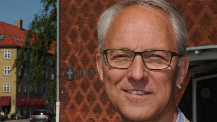 Mand med briller og blå skjorte står foran rødt murværk med "ADEGA" skilt.