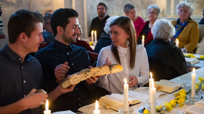 En gruppe af mennesker sidder ved et bord med lysstake og blomster, mens en mand holder en lang brødstang.