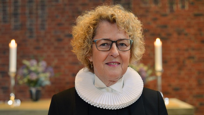 En kvinde med hårbånd og briller står foran murvæg med lysstake og blomster.