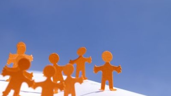 Fünf orange Figuren stehen im Halbkreis auf einem weißen Hügel vor blauem Himmel.