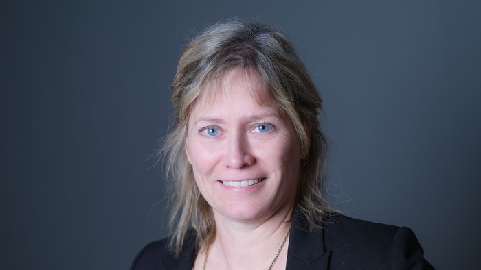 En kvinde med blondt hår og blå øjne, der smiler og er tagen i en sort jakke.