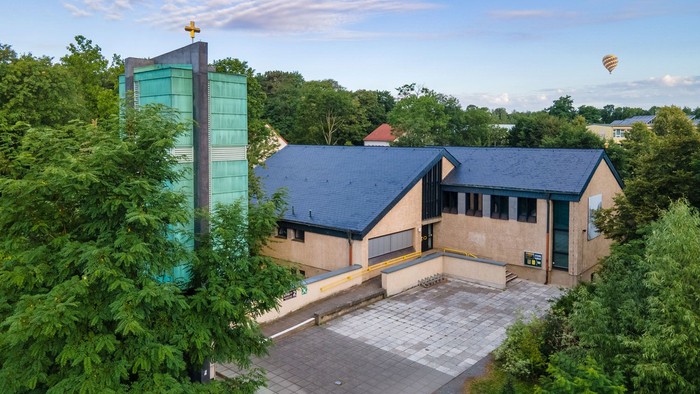 Ein modernes Gebäude mit Glasfassade und Flachdach, umgeben von Grünflächen und Bäumen, mit einem Heißluftballon im Hintergrund.