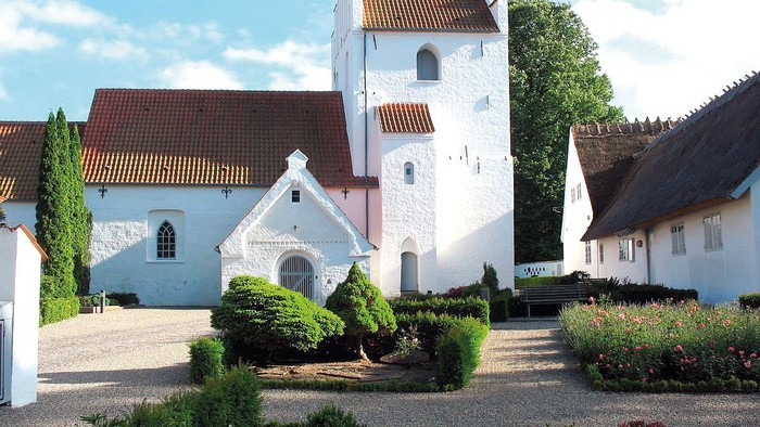 En smuk og hvid kirke med tårn og røde tag i en trædit og grønne omgivelser
