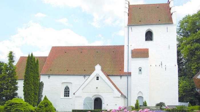 Et hvidt kirkebygning med røde tag og en tårn, omgivet af grønne planter og blomster.