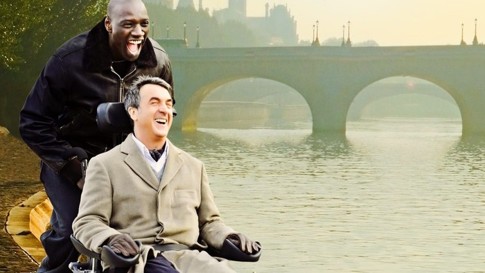 En mand i håndstol og en andet mand ved en flod med bro i baggrunden