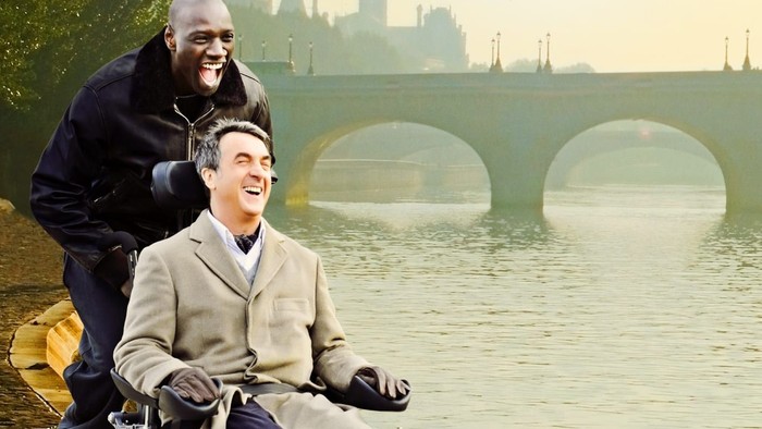 En mand i håndstol og en andet mand ved en flod med bro i baggrunden