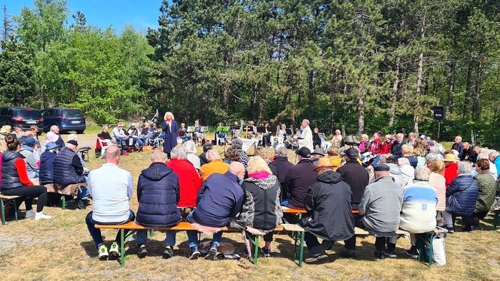 Große Menschenmenge sitzt im Freien auf Stühlen und hört einem Redner zu, umgeben von Bäumen.